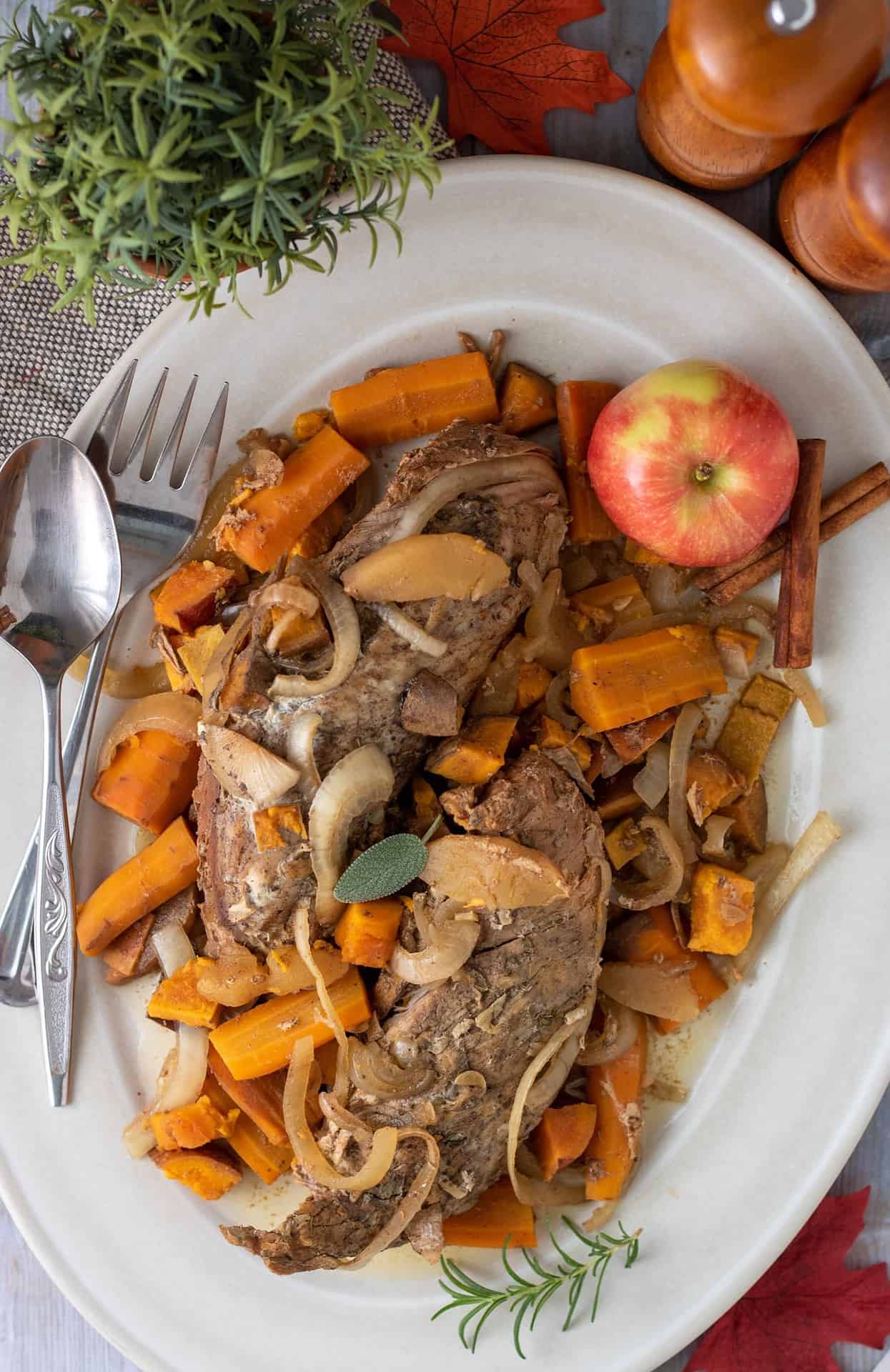 A large oval serving platter with apple cider pork tenderloin on top of carrots, onions and sweet potatoes.  There's a red apple on the serving platter and two silver serving utensils.  There's a small fresh rosemary plant in the background