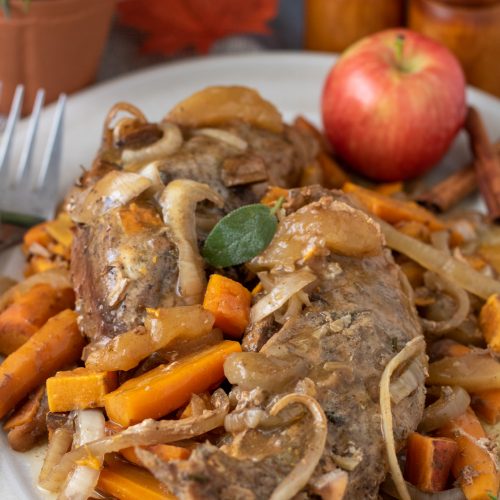 A oval white serving plater topped with pork tenderloin with onions, carrots, sweet potatoes and apples. There's a large silver serving utensil next to the pork with a red apple on the platter. There's a small rosemary plant in the background and red maple leaf