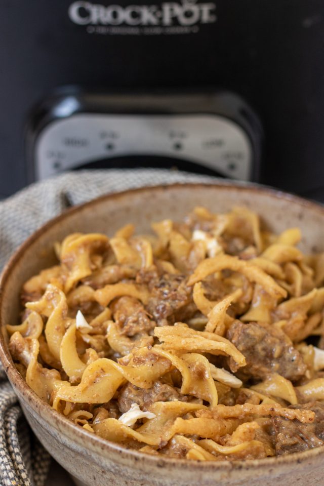 A brown specked bowl with egg noodles and beef stew meat. It's creamy and there's a black crockpot in the background.