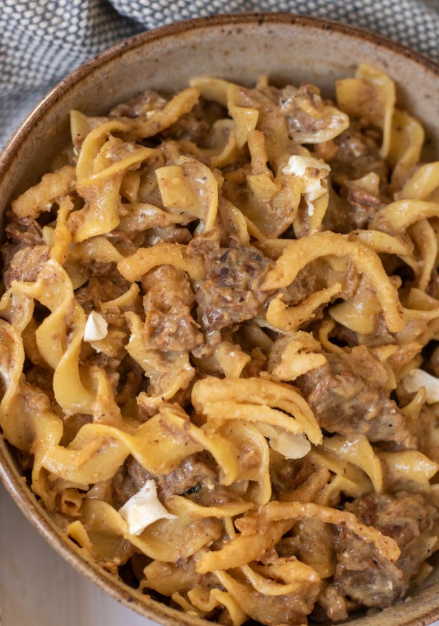 A brown speckled bowl full of egg noodles topped with french onion beef stroganoff. You can see the chunks of cream cheese and the noodles are creamy and buttery.