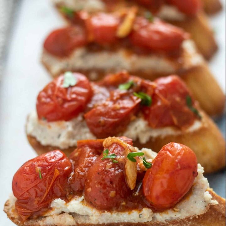 A marble serving board with slices of bruschetta that’s topped with roasted tomatoes & garlic on top of a ricotta & bean purée. They’re sprinkled with fresh parsley