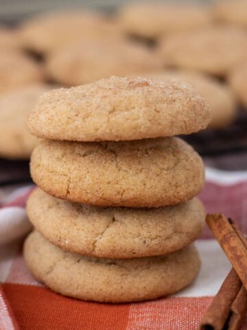 4 snickerdoodle cookies stacked on top of each other. They're sitting on a orange plaid kitchen towel, with two cinnamon sticks next to them. There's a cooling rack with more cookies in the background