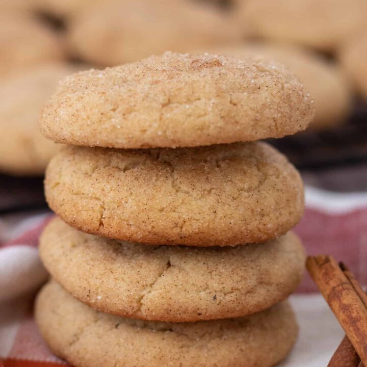 4 snickerdoodle cookies stacked on top of each other. They're sitting on a orange plaid kitchen towel, with two cinnamon sticks next to them. There's a cooling rack with more cookies in the background