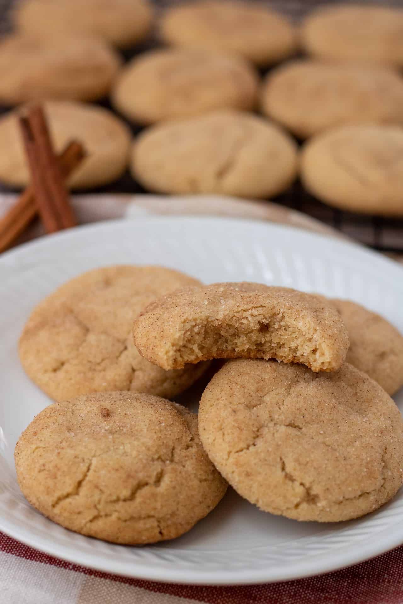A small round white plate with 5 chai spice snickerdoodle cookies. The top cookie has a bite taken out of it. There's two cinnamon sticks with cooling rack of more cookies in the background