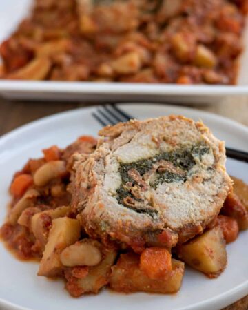 A white dinner plate with a slice of pork roast that's stuffed with spinach. It's on top of a tomato sauce with diced potatoes, carrots and cannellini beans. In the background there's a white rectangle serving platter with a pork loin roast