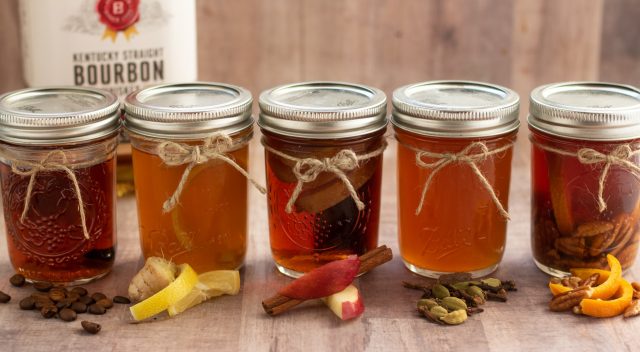 5 small mason jars with infused whiskey. The ingredients are in front of each jar like whole coffee beans, lemon and ginger, apple slice and a cinnamon stick. There's a handle of bourbon in the background.