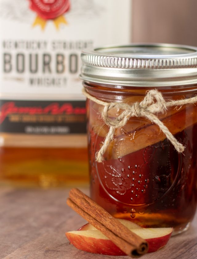A small mason jar on a wooden surface that's been infused with apple and cinnamon. There's a hemp string tied around the jar and a whole apple slice with cinnamon stick in front of it. There's a bottle of bourbon in the background