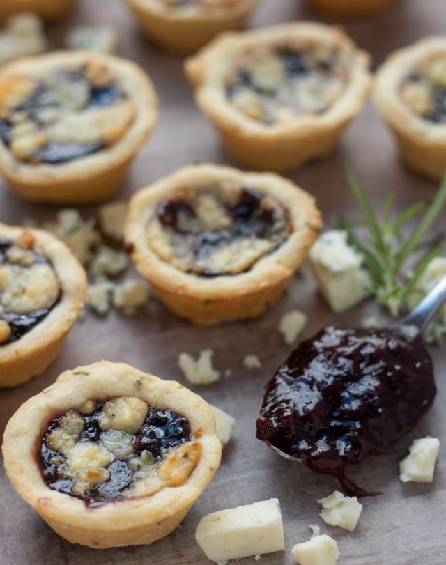 Several mini shortbread tarts that are filled with jam and topped with cheese. There's gorgonzola crumbles sprinkled around the tarts and a spoonful of blackberry jam in the background with a fresh rosemary sprig