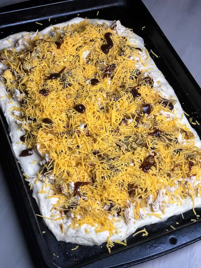 A baking sheet of pizza dough stretched to a 13” x 7” rectangle and topped with barbecue sauce, shredded chicken, caramelized onions and shredded cheese.