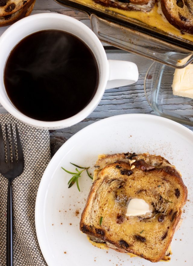 A small round white plate with Monte Cristo french toast casserole on it. There's a black fork on a grey specked dinner napkin next to the dish with a white coffee mug filled with black coffee and a dish of butter