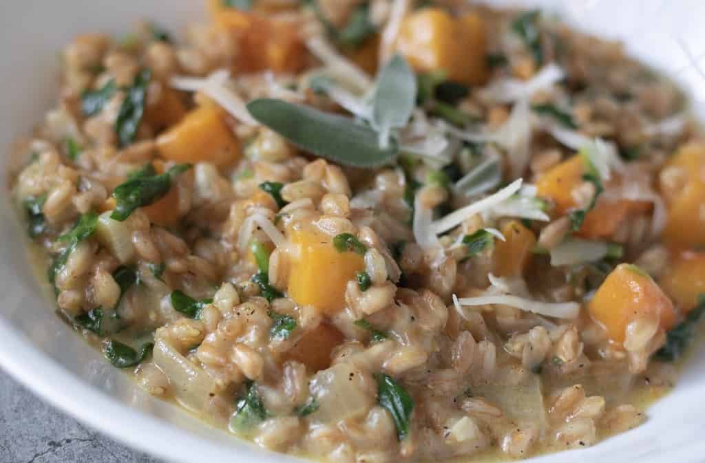 A closeup shot of a white bowl filled with farro risotto. It has pieces of butternut squash and spinach and topped with Parmesan cheese and fresh sage leaves