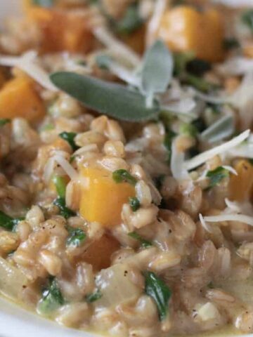 A closeup shot of a white bowl filled with farro risotto. It has pieces of butternut squash and spinach and topped with Parmesan cheese and fresh sage leaves