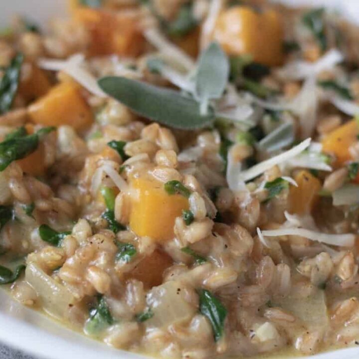 A closeup shot of a white bowl filled with farro risotto. It has pieces of butternut squash and spinach and topped with Parmesan cheese and fresh sage leaves