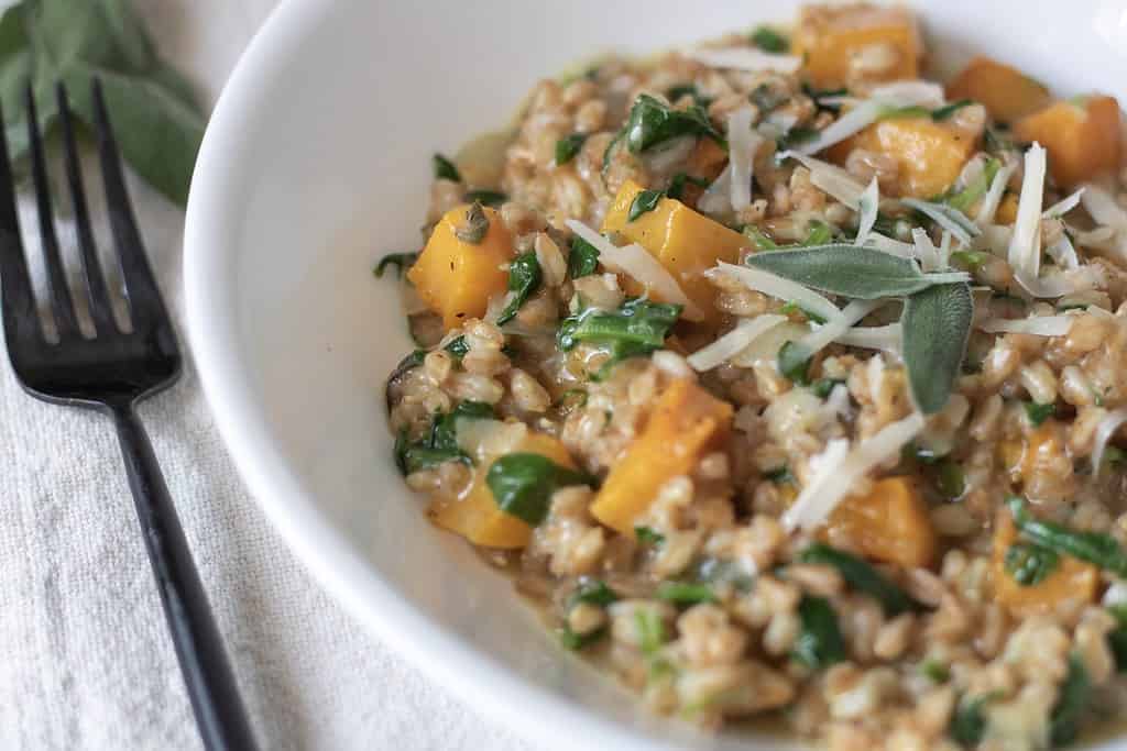 A white bowl filled with creamy farro risotto with butternut squash and grated Parmesan cheese. There’s a fork next to the bowl and a piece of fresh sage leaves in the background.