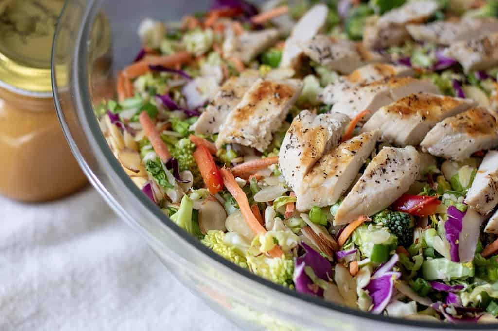 A big bowl of chopped Asian salad with grilled chicken, purple cabbage, broccoli, cucumber and lettuce. Topped with almonds and sesame seeds. A jar of easy Asian orange sesame peanut dressing is next to the salad bowl.
