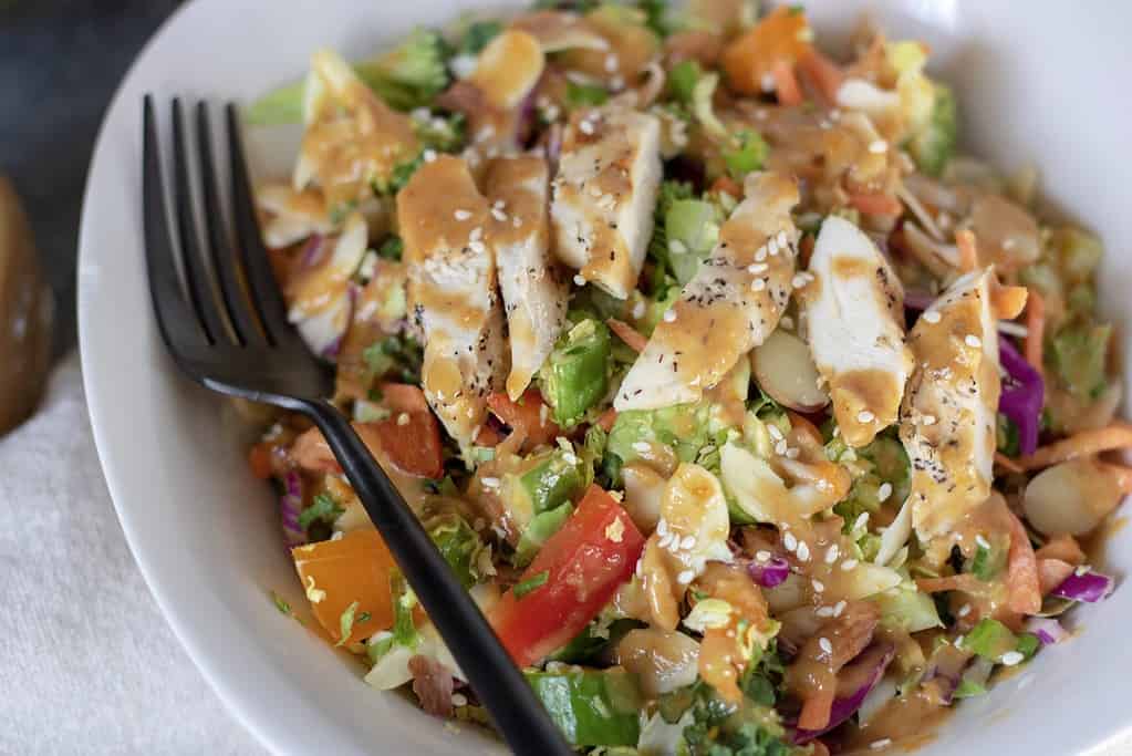 A white bowl filled with crunchy Asian salad with red peppers, cabbage, almonds, broccoli and lettuce. It’s drizzled with a delicious orange sesame peanut dressing and topped with slivered almonds and sesame seeds. It’s easy, healthy and low carb.