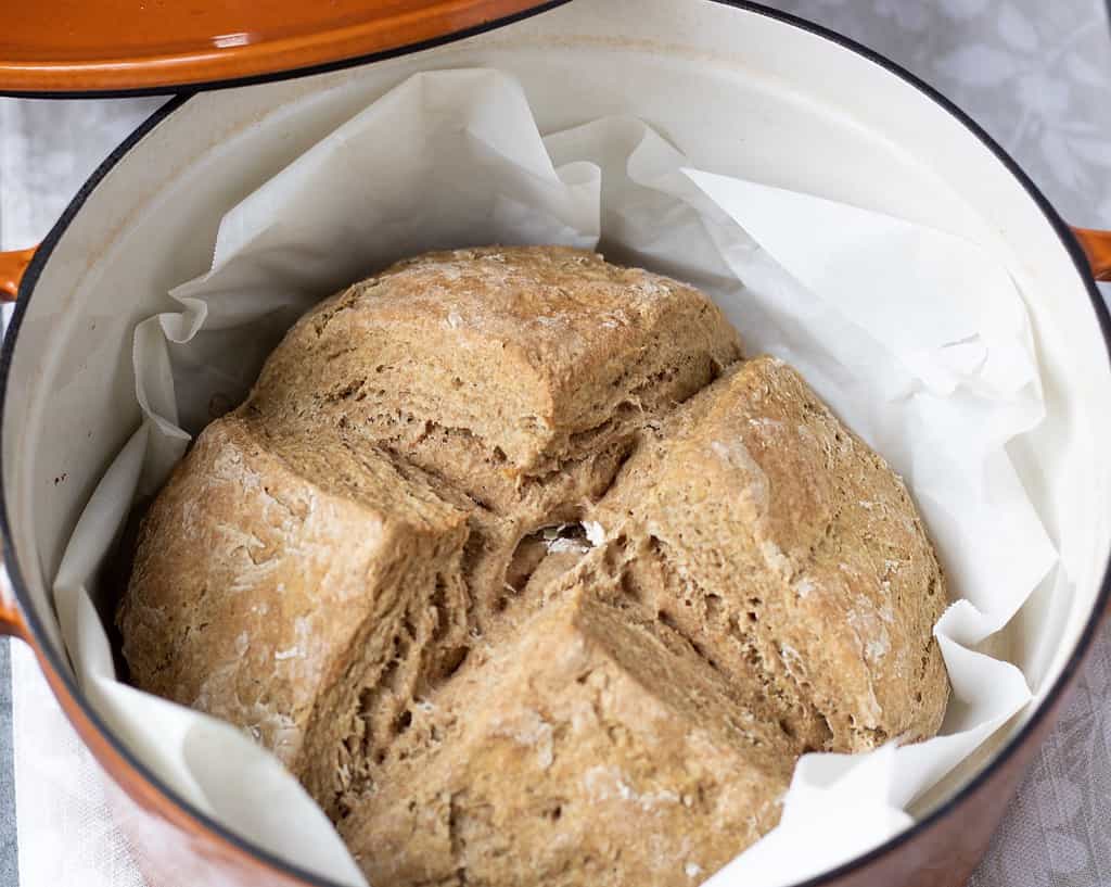 A loaf of Irish soda bread on top of parchment paper in an orange Dutch oven pan. It was scored with an x before baking in the oven.