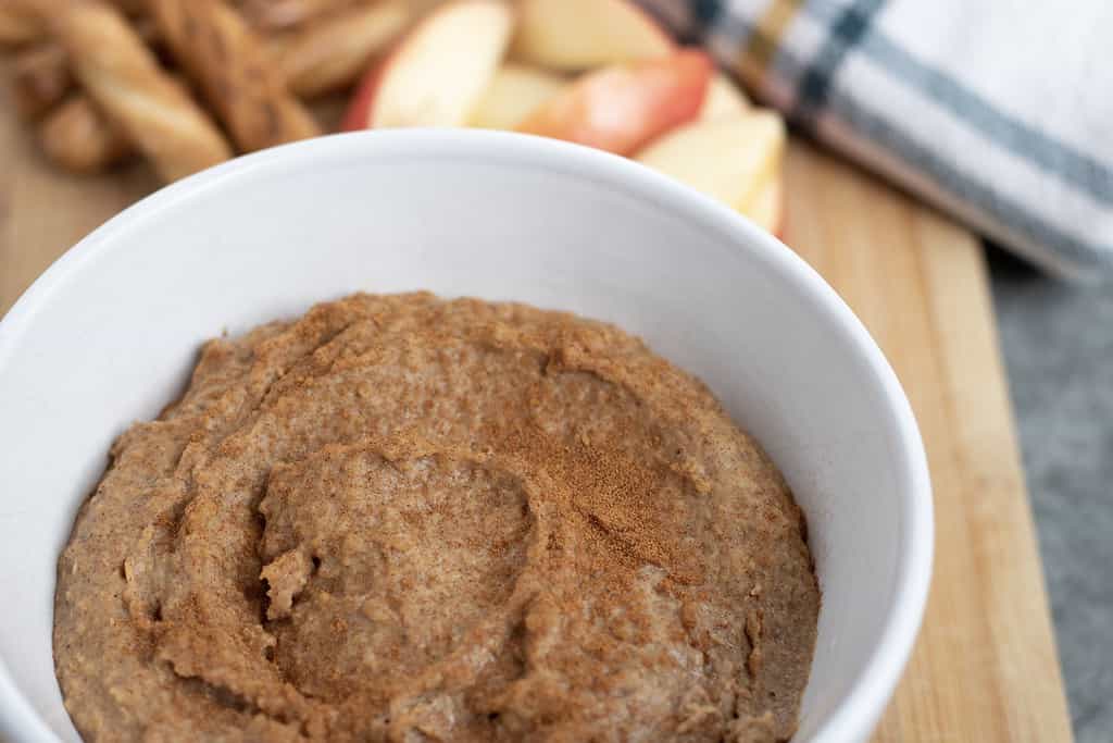 A white bowl full of Snickerdoodle Dessert Hummus, sprinkled with cinnamon. Honey wheat pretzel sticks and red apple slices are in the background.