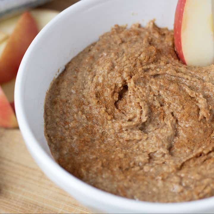 A bowl with Snickerdoodle Dessert Hummus with a slice of apple that’s being dipped in the bowl. Pretzel sticks are in the background for this easy and healthy snack recipe
