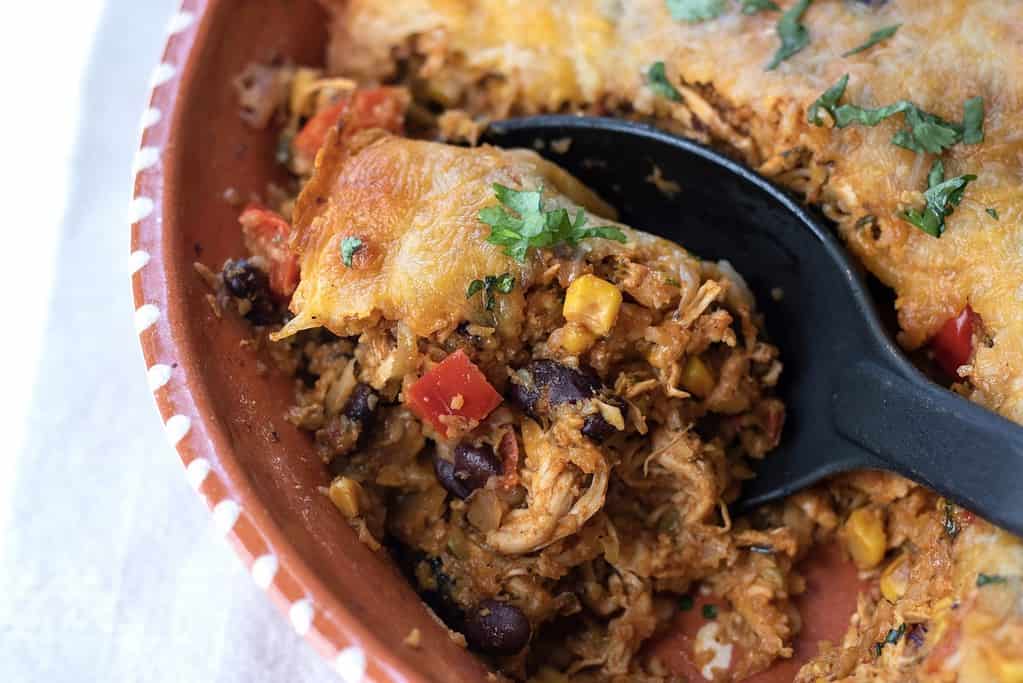 A casserole dish with low carb Mexican casserole with a serving spoon scoop. It has shredded chicken, taco seasoning, black beans, corn and red peppers for an easy and healthy dinner.