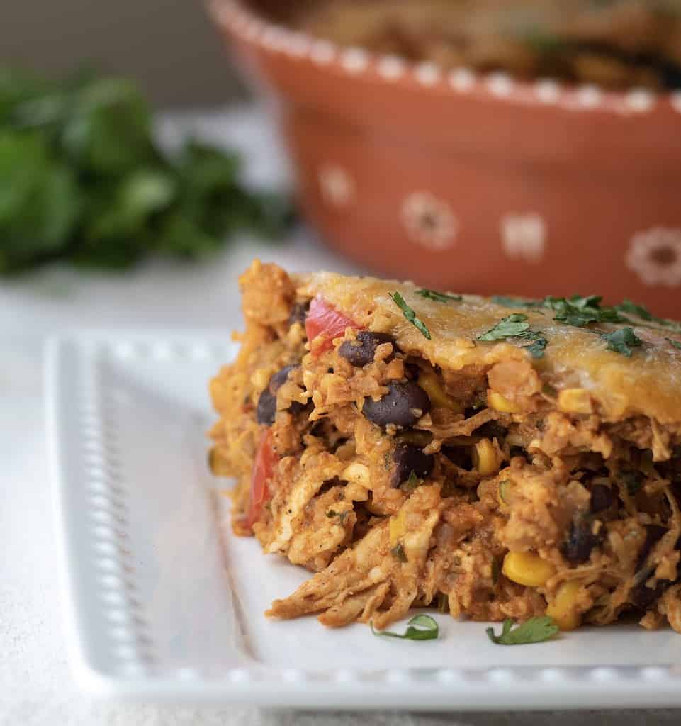 A white square dish with a serving of Mexican cauliflower rice casserole with shredded chicken and cheese. A round casserole dish is in the background with a bunch of fresh green cilantro