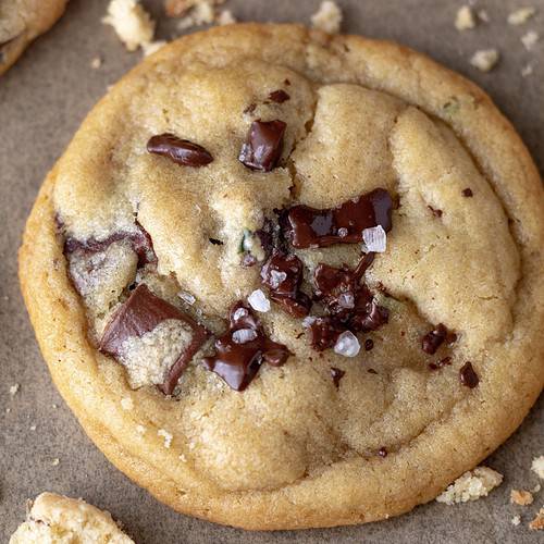 A perfectly crisp and gooey cookie made with chocolate chunks and lavender. It’s on brown parchment paper and the chocolate chunks are slightly melted and there’s a sprinkle of sea salt on top. There are cookie crumbs on the baking sheet surrounding the cookie.