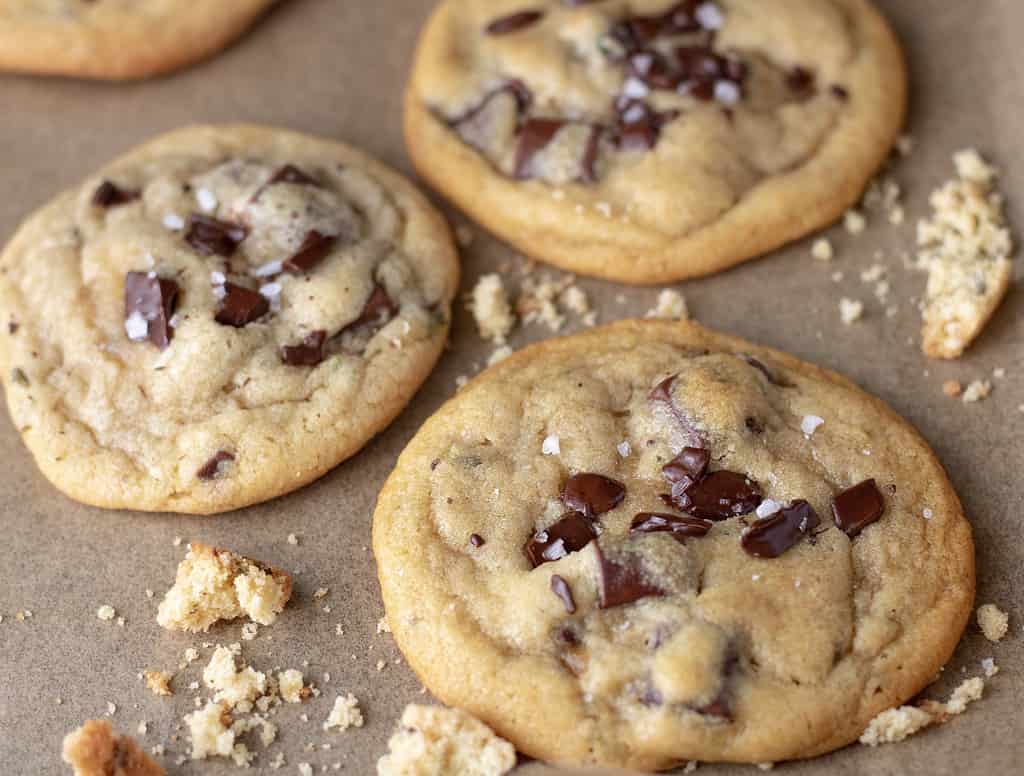 Three perfectly crisp and gooey cookies made with chocolate chunks and lavender. They’re on brown parchment paper and the chocolate chunks are slightly melted and there’s a sprinkle of sea salt on top. There are cookie crumbs on the baking sheet surrounding the cookies.