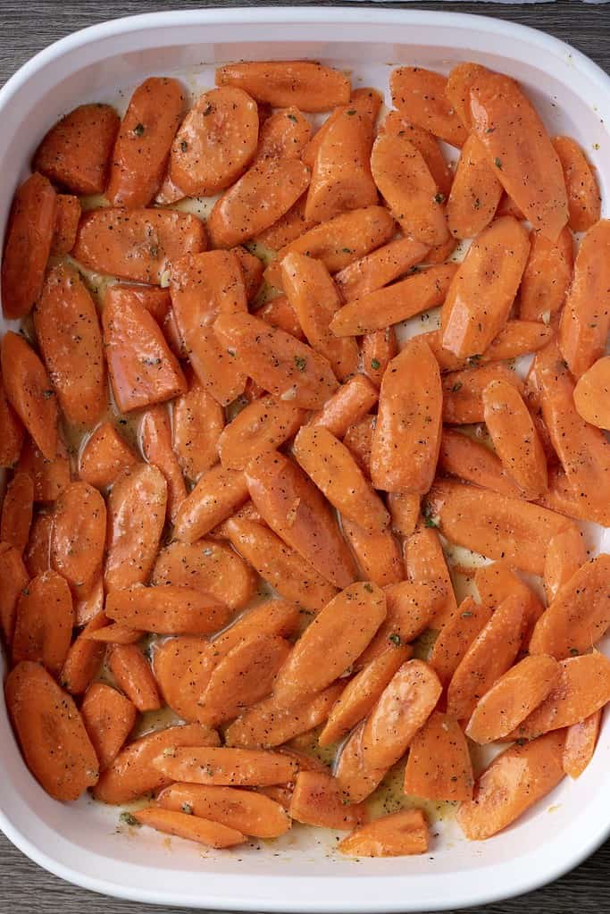A white casserole dish of thin diagonally sliced carrots tossed in olive oil, garlic, thyme, lemon, maple syrup and ginger.