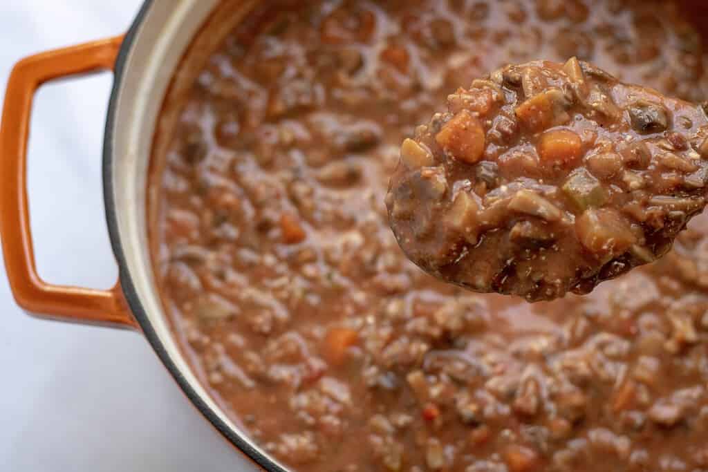 An orange Dutch oven filled with mushroom and eggplant bolognese showing a serving spoon closeup shot. Its an vegetarian version of the traditional Italian recipe.