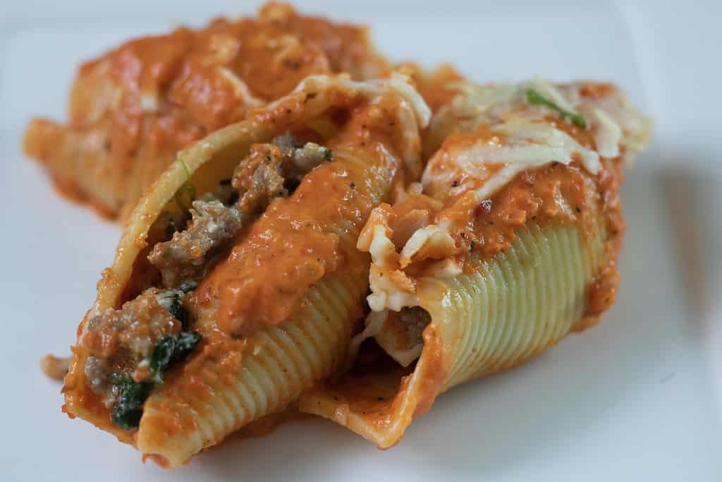 3 stuffed shells on a white plate covered in melted mozzarella cheese and filled with ricotta, sausage, spinach and goat cheese. They’re topped with a roasted red pepper cream sauce.