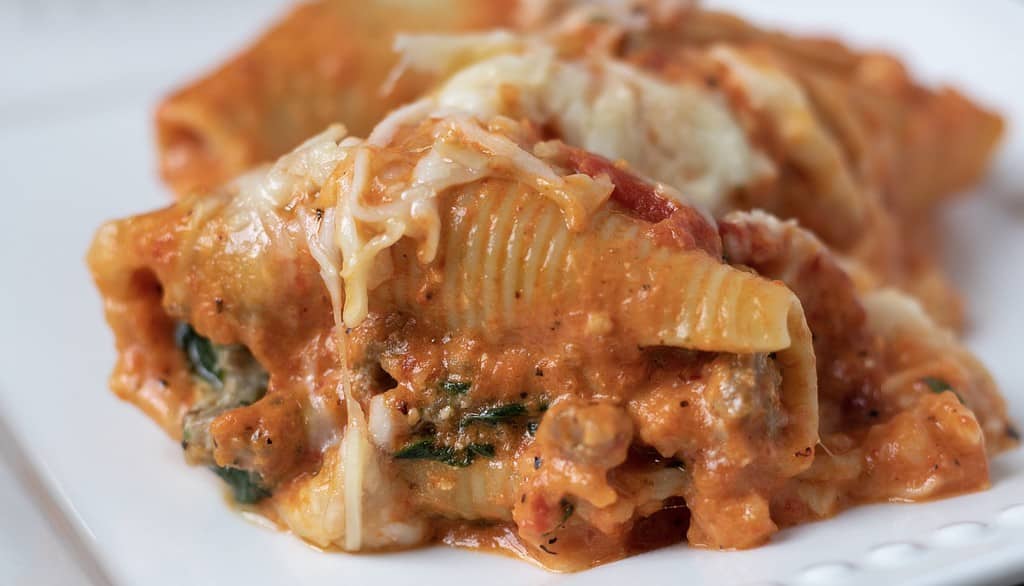 A white square dish with 3 stuffed shells that are covered in a creamy red peppers sauce and melted mozzarella cheese.