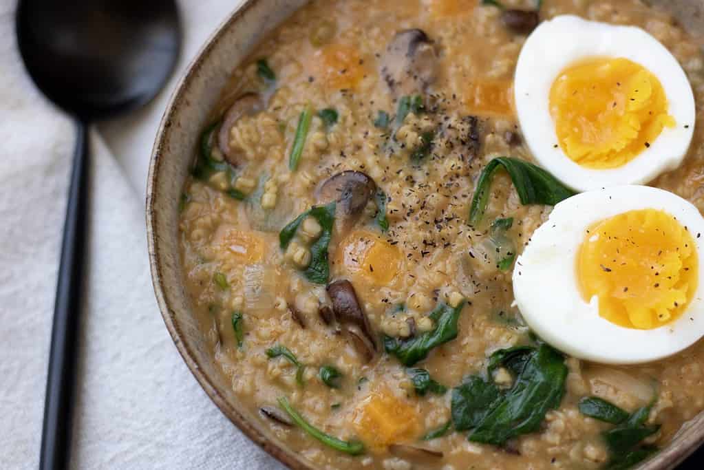 A brown speckled bowl filled with cheesy oatmeal made from steel oats. It’s cooked with spinach and mushrooms and topped with a boiled egg.