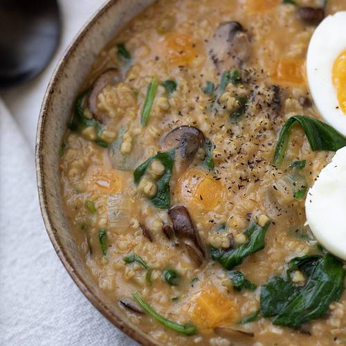 A brown speckled bowl filled with cheesy oatmeal made from steel oats. It’s cooked with spinach and mushrooms and topped with a boiled egg.