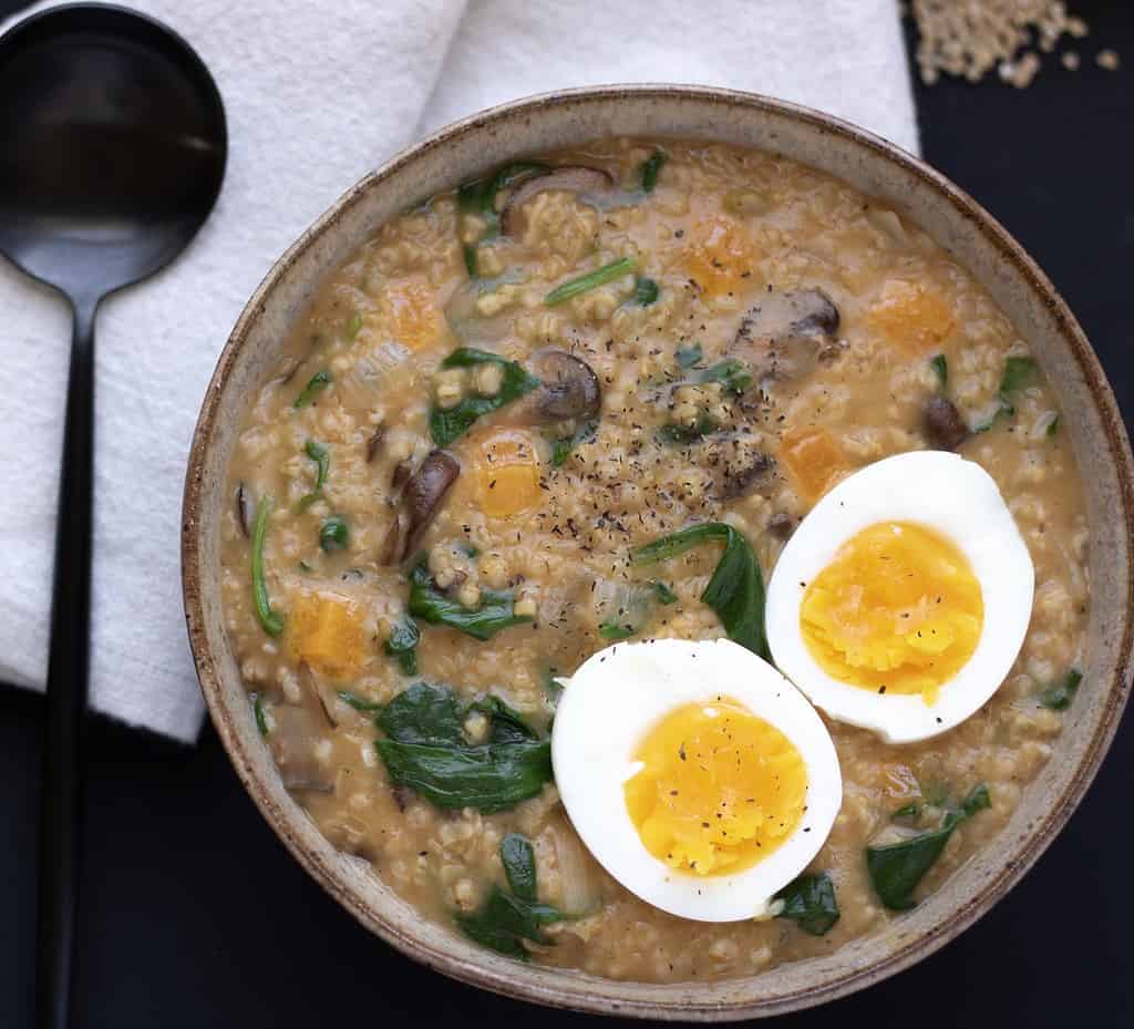 A bowl of savory oatmeal with spinach and mushrooms. A soft boil egg is cut in half and placed on top of the oatmeal. Raw steel oats are spilled in the background.