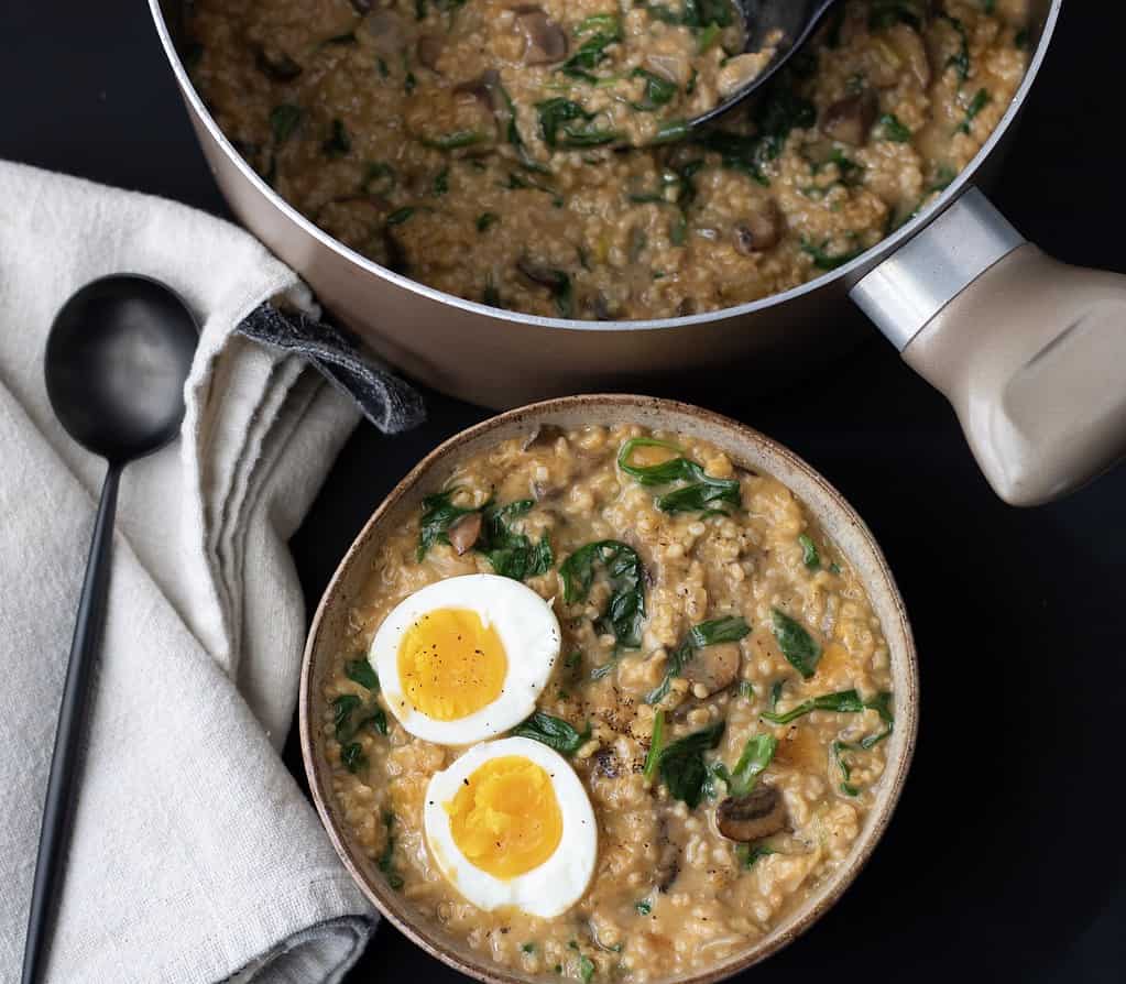 An overhead shot of a bowl of savory oatmeal. There’s a round pot of oatmeal in the background and a beige napkin with black spoon. There’s spinach, mushroom and cheddar in the oatmeal.