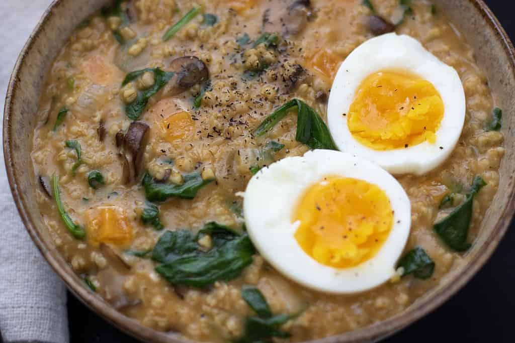 A bowl of creamy oatmeal with spinach, mushrooms and cheddar cheese. A soft boiled egg is cut in half and served on top with a sprinkle of black pepper