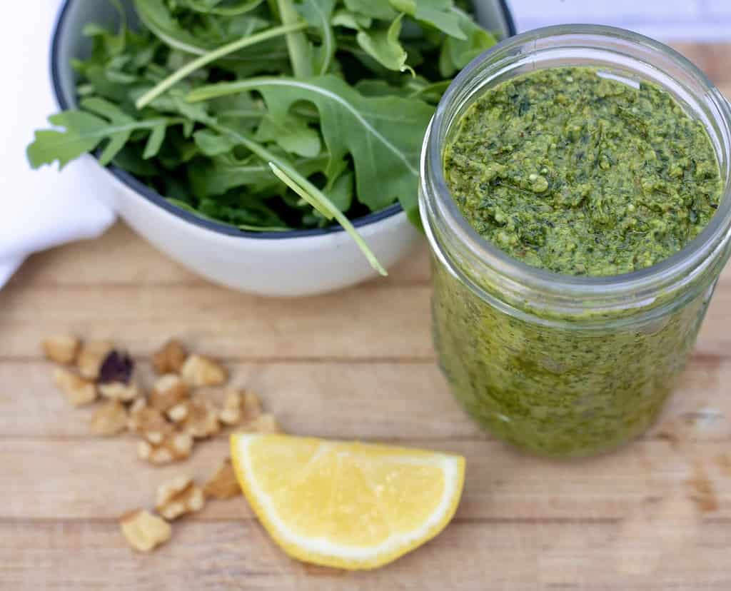 A jar of homemade pesto that’s made with arugula, spinach, basil and walnuts. There’s a lemon wedge, walnut pieces and a small bowl full of fresh arugula in the background