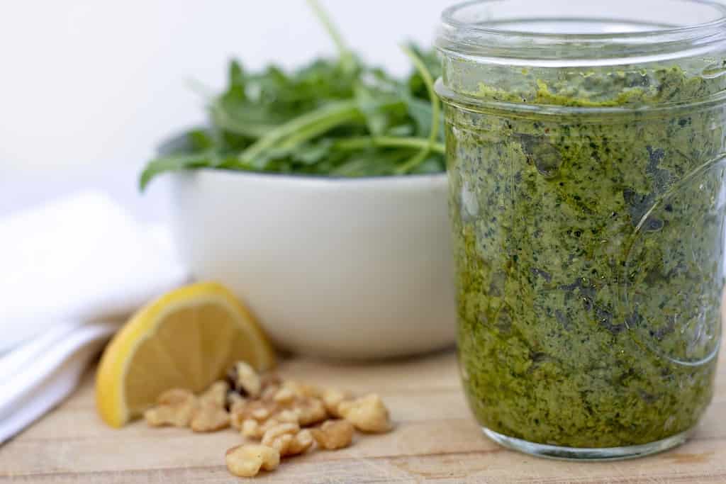 A head on shot of a small mason jar filled with homemade pesto sauce. It’s quick and easy to make using arugula basil and walnuts. There’s a slice of lemon next to the jar of pesto and fresh arugula leaves in the background