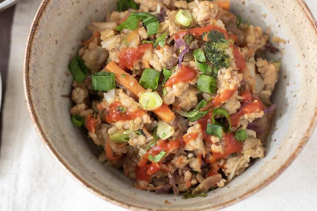 A bowl full cauliflower rice stir fry with broccoli, cabbage and carrots. It’s made in a homemade teriyaki sauce and finished with sliced scallions and sriracha for an easy and healthy lunch or dinner