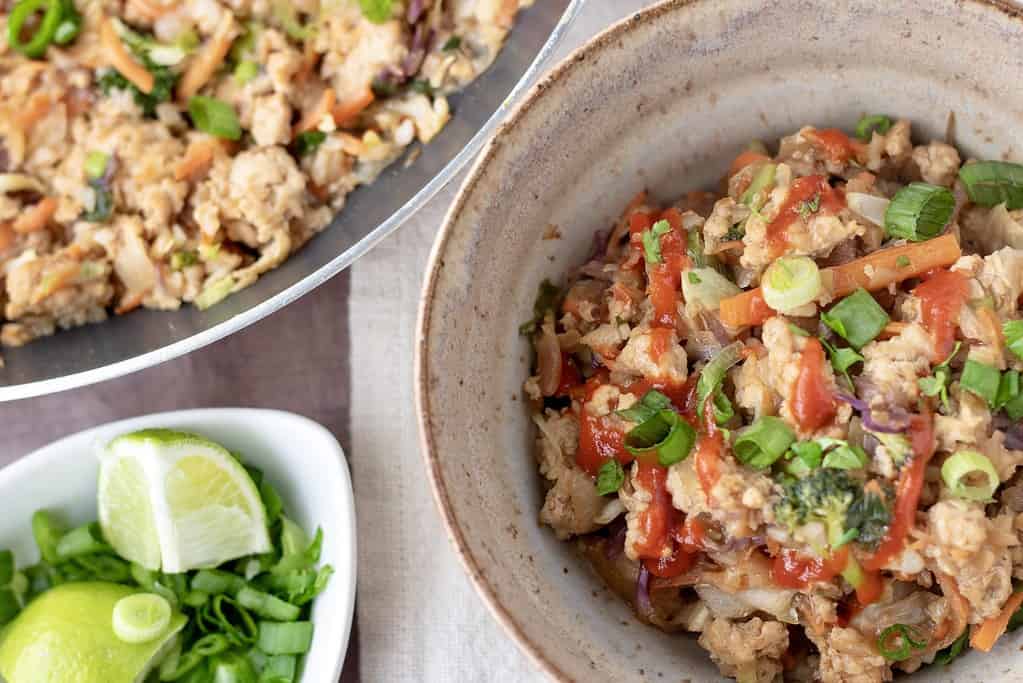 A brown speckled bowl filled with Asian stir fry cauliflower rice. It’s garnished with scallions and sriracha sauce. There’s a skillet pan in the background and a small bowl of lime wedges.