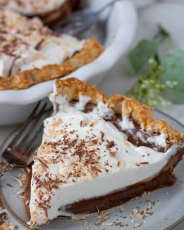 A small pie plate with a slice of chocolate coconut cream pie. There's a fork next to the plate and a white pie plate with coconut pie in the background. The pie slice is topped with whipped cream, toasted coconut and shaved chocolate pieces.