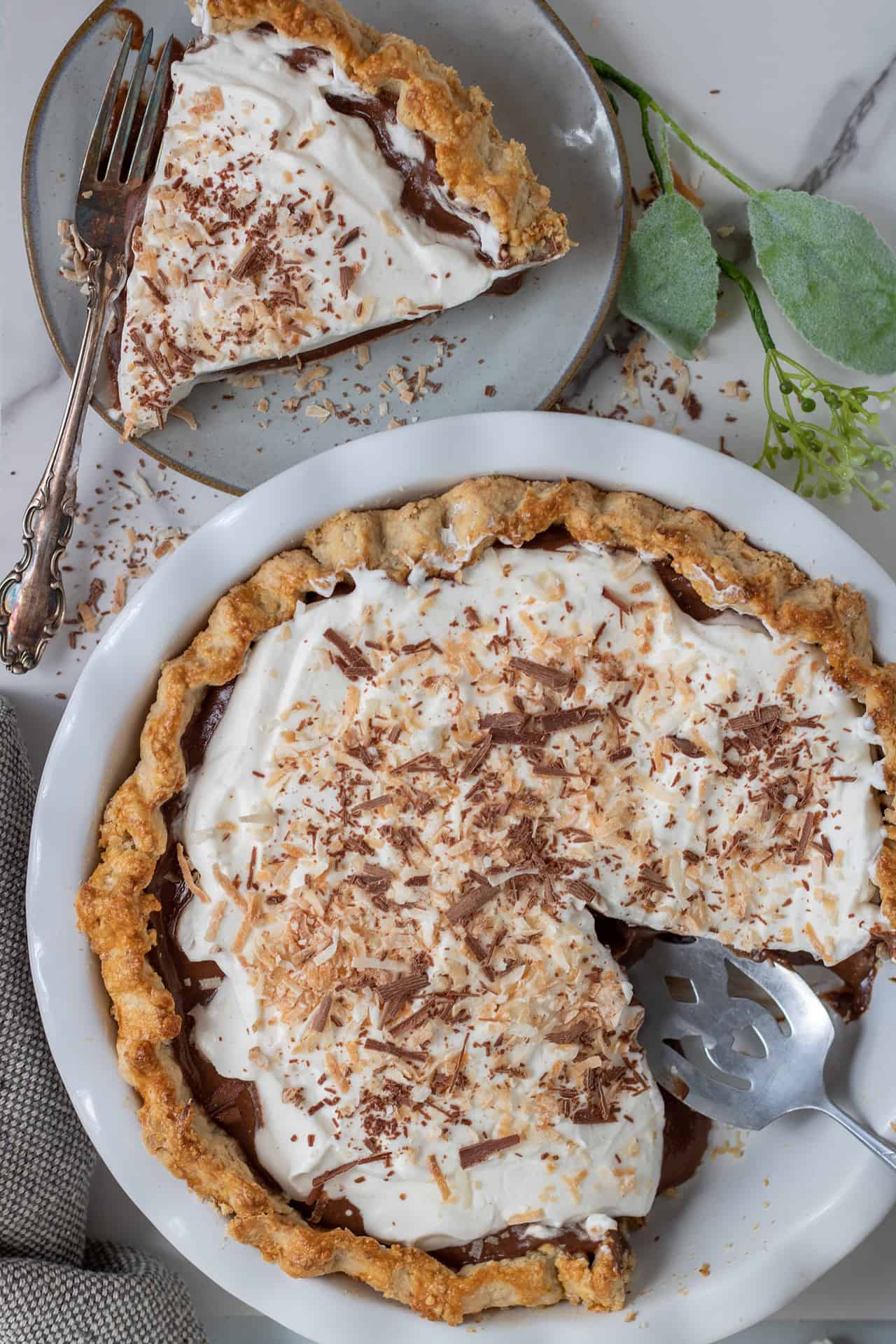 A white pie plate filled with chocolate coconut cream pie. There's toasted coconut and chocolate shavings on top. There's a small plate in the background with a slice of pie on it. There's a green flower in the background.