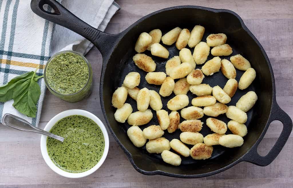 A cast iron pan with cauliflower gnocchi that’s been pan fried until golden and crispy. A bowl of creamy pesto is set next to it, with some fresh basil leaves