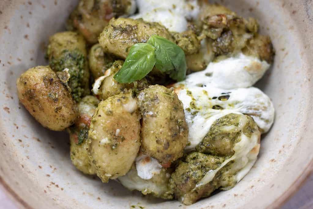 A brown speckled bowl full of homemade crispy & golden cauliflower gnocchi that’s covered with creamy pesto sauce and melted fresh mozzarella cheese. A piece of fresh basil is on top of the gnocchi bake.
