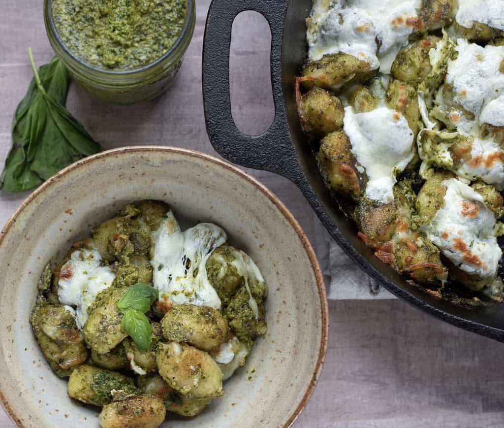 A bowl full of cheesy cauliflower gnocchi with pesto with a cast iron pan of the crispy gnocchi set next to it. They’re covered with fresh mozzarella cheese and fresh basil. The mozzarella is melted until slightly golden and a jar of the homemade pesto is in the background