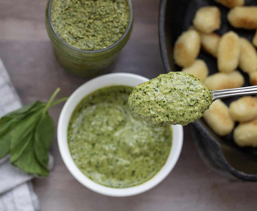 A spoonful of creamy pesto sauce. There’s a jar of the homemade pesto in the background and a cast iron pan of crispy cauliflower gnocchi