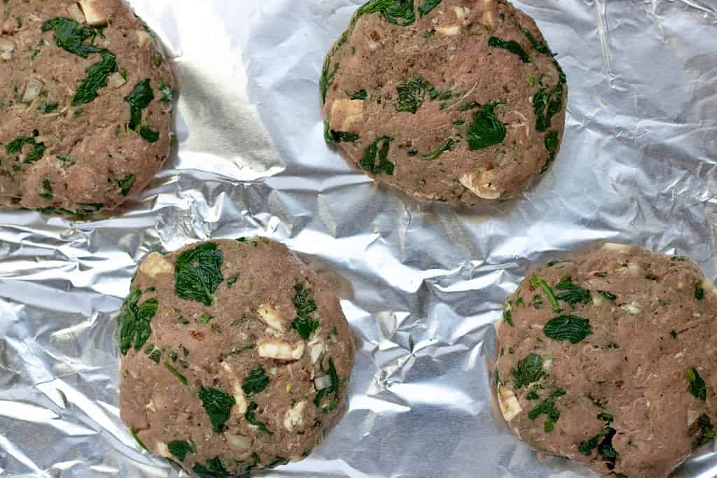A sheet pan lined with aluminum with raw turkey burger patties. They’re stuffed with spinach and mozzarella and baked in the oven for a healthy and juicy burger