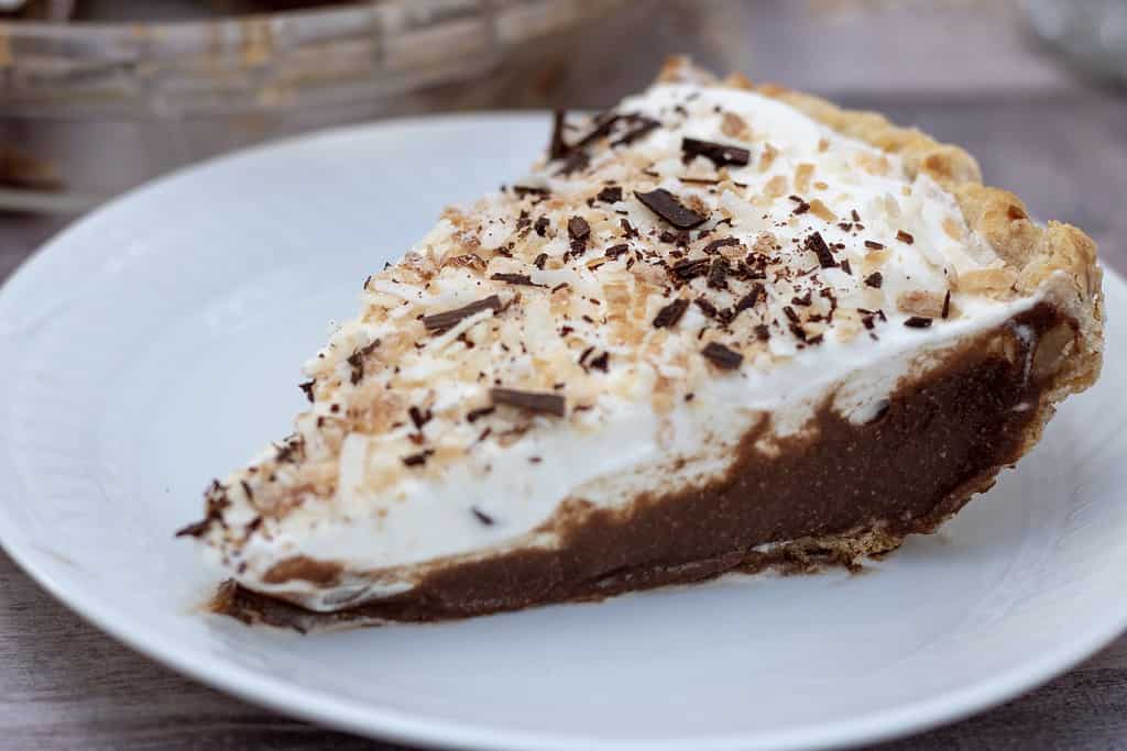 A slice of chocolate coconut cream pie in a white plate. There’s toasted coconut and shaved chocolate sprinkled on the top