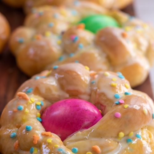 3 loaves of easter bread on a wooden surface. They're glazed with a sugar coating with easter sprinkles. There's a dyed egg in the middle of each loaf.