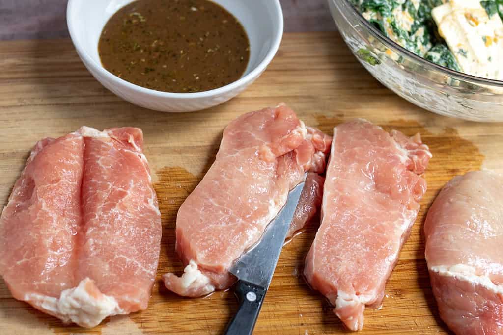 A wooden cutting board with 3 boneless pork chops that are being cut sideways so they can be filled with a spinach and cheese stuffing. A bowl of the spinach and cheese filling is in the background as well as a small white bowl of honey dijon balsamic glaze.